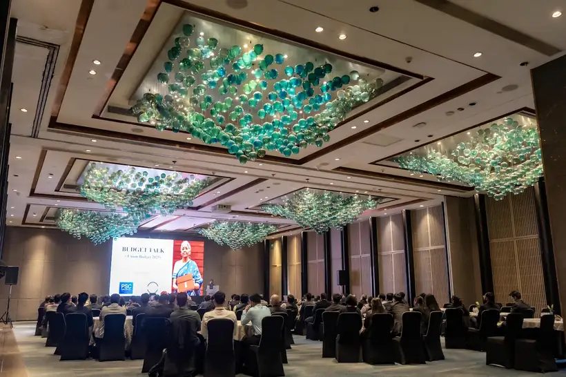 A seated audience in a banquet hall facing a lit stage and screen