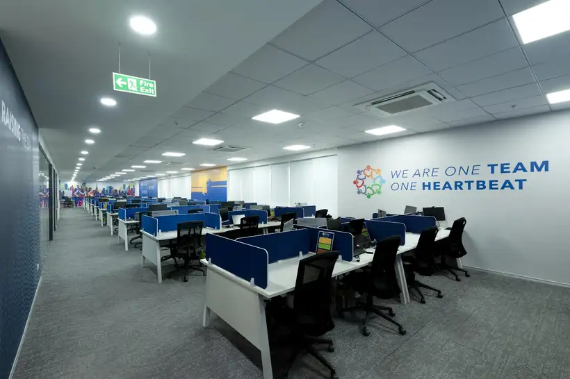 A long open-plan office floor with blue partitions and a wall graphic reading we are one team one heartbeat